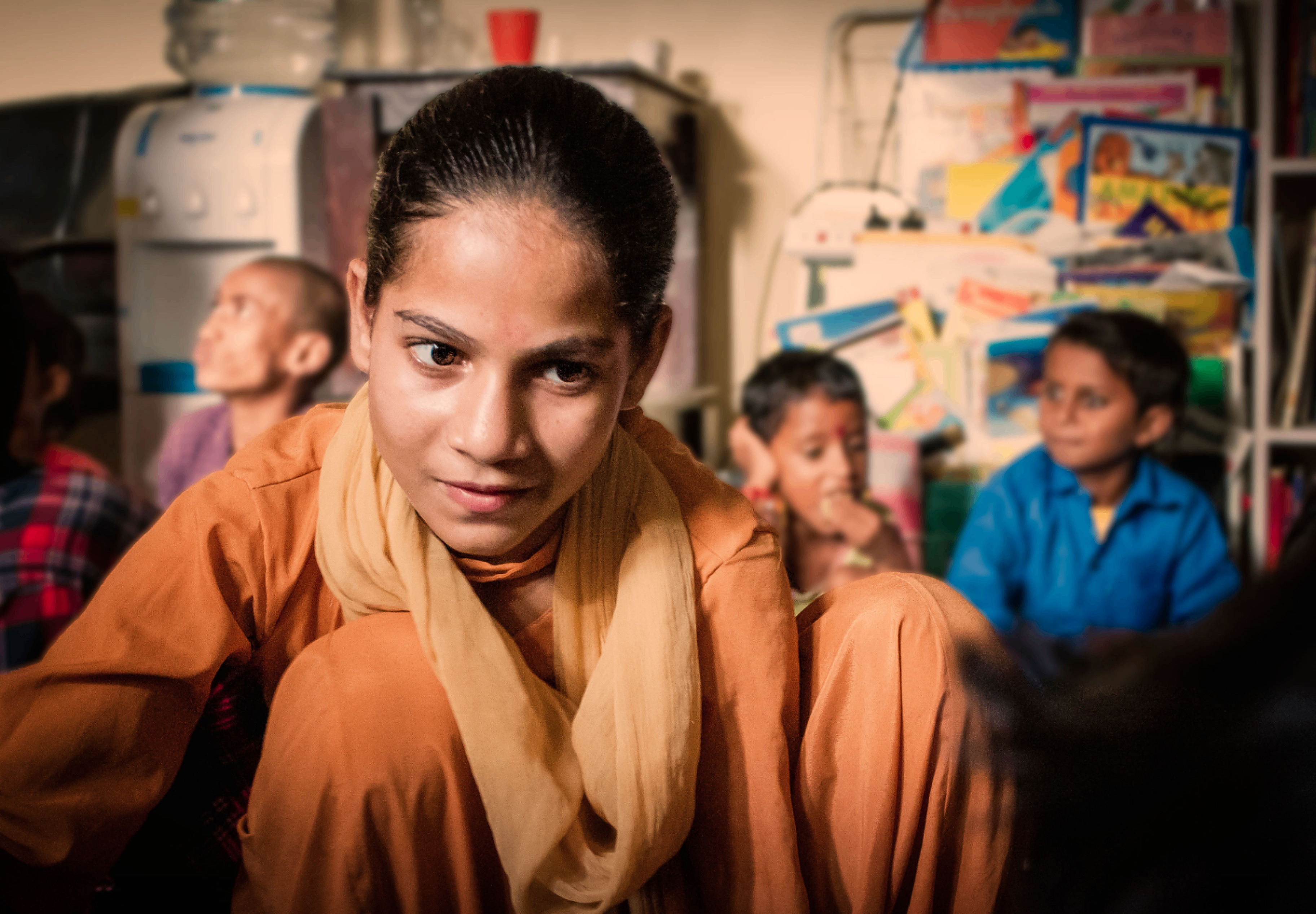 A girl in an orange kurta inside a learning space surrounded by books, drawings and other children